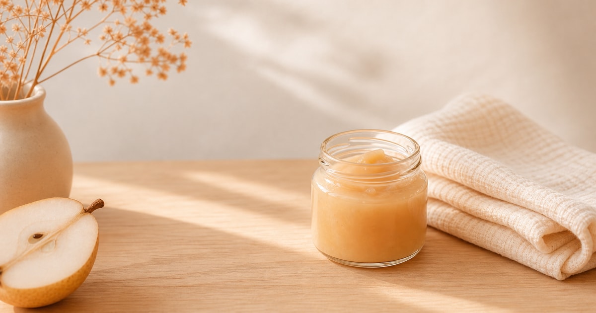 A small glass jar of pureed pear next to a folded muslin cloth resting on a wooden surface in soft morning light