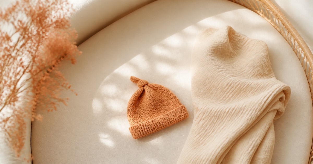 A knitted newborn hat and swaddle blanket on a bassinet sheet in soft morning light