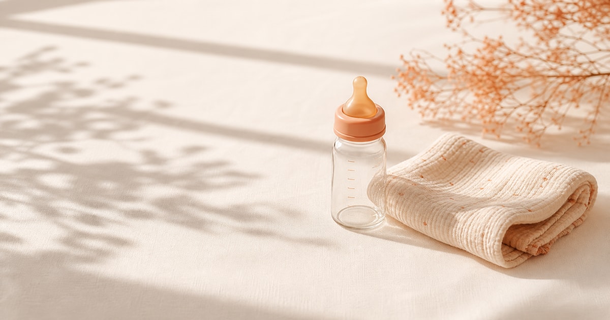 A small baby bottle and folded muslin cloth on a cream linen surface